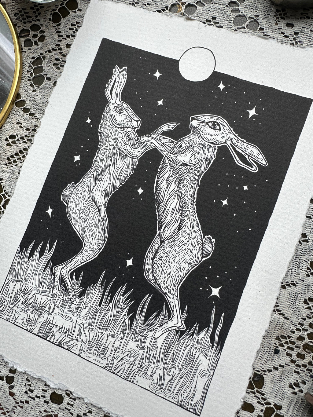Black and white illustration of two hares on a starry night sky background, placed on a textured fabric surface.
